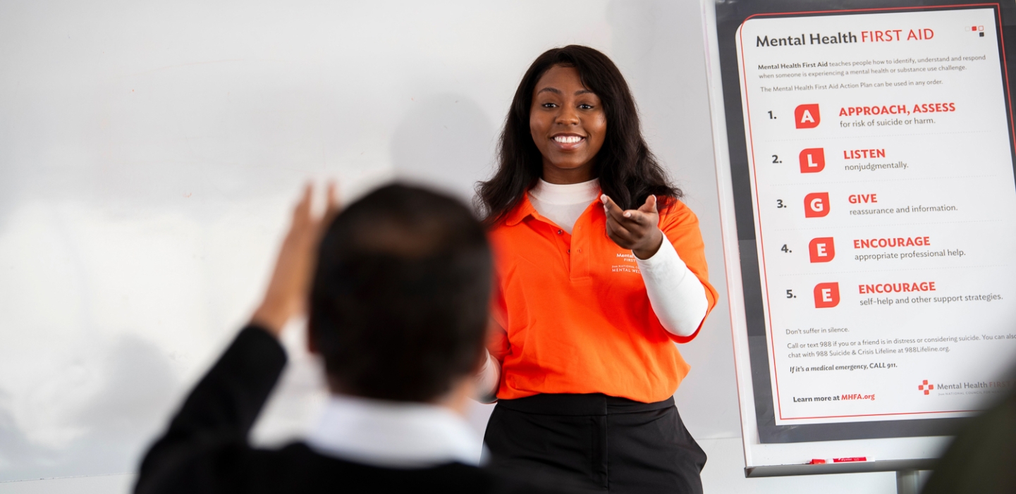 Female instructor stands in front of a Mental Health First Aide poster and answers an attendee question.