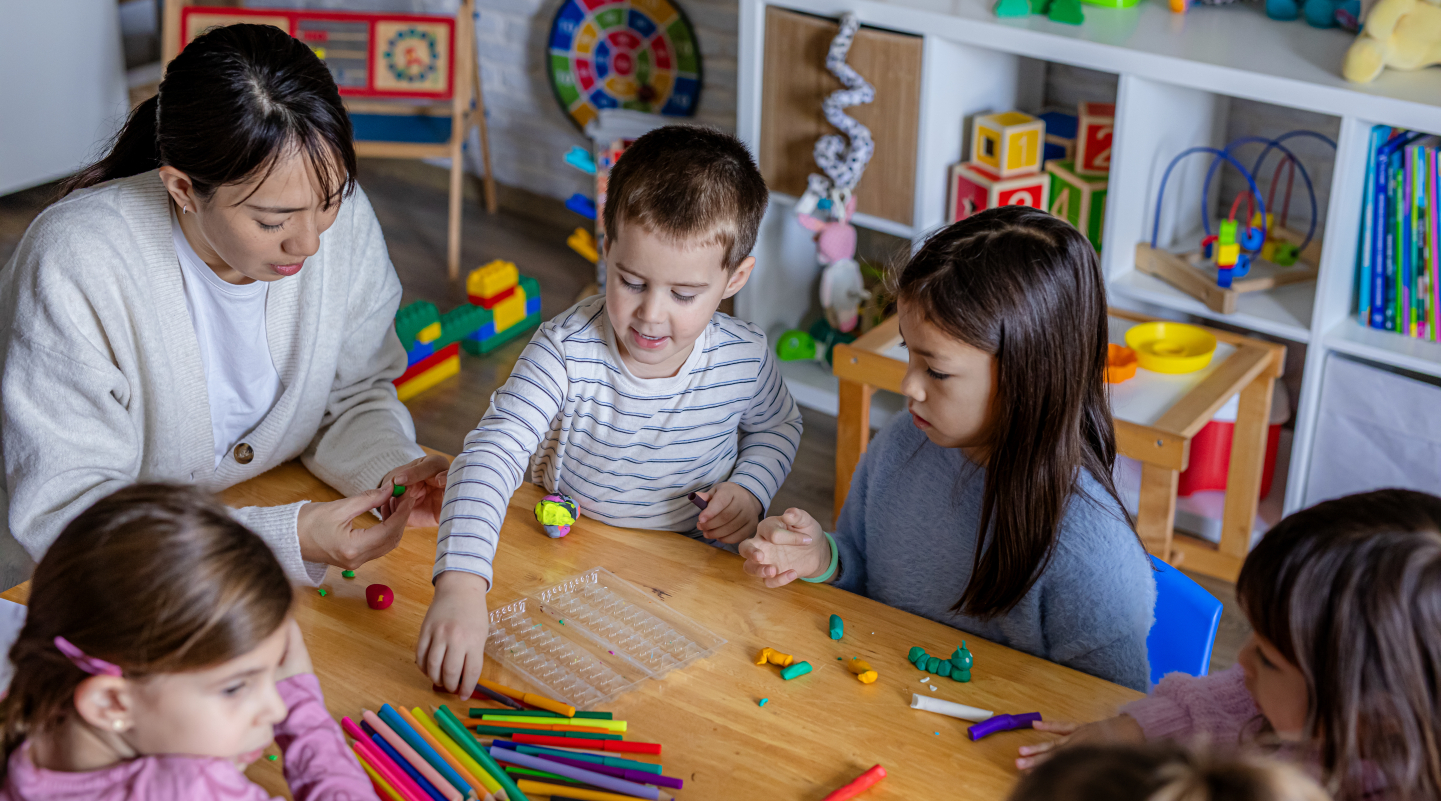 A photograph of a childcare educator with several children around a low table, playing with various craft supplies.