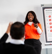 Female instructor stands in front of a Mental Health First Aide poster and answers an attendee question.