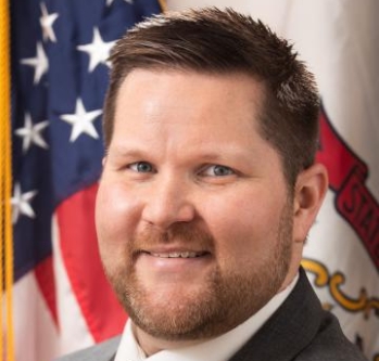 Federal official with brown hair smiling in front of the U.S. flag