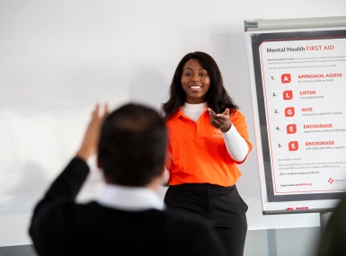 Female instructor stands in front of a Mental Health First Aide poster and answers an attendee question.