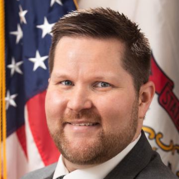 Federal official with brown hair smiling in front of the U.S. flag