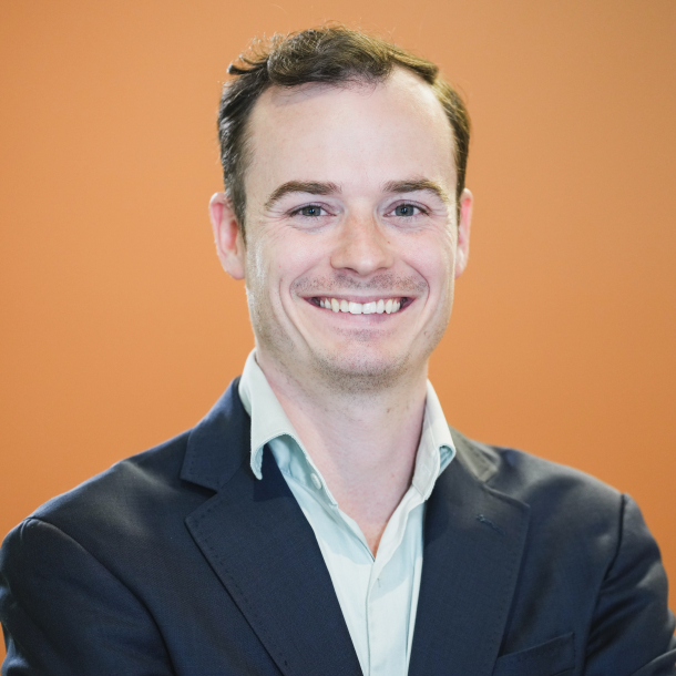 Picture of Seamus Dowdall smiling in a suit jacket and button down with orange background
